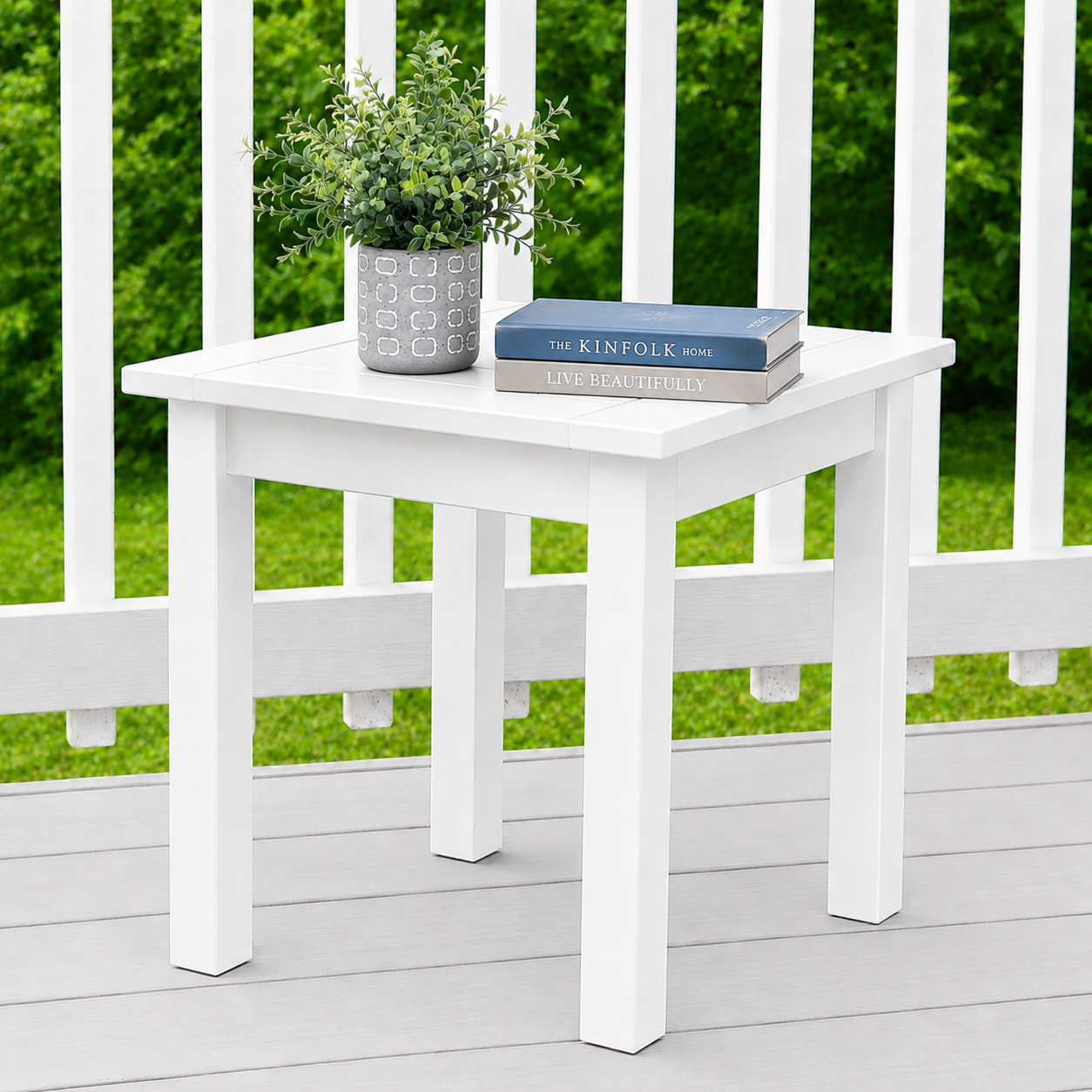 A white square side table on a light gray deck with a white railing, featuring a potted green plant and two books on top, set against a background of lush green foliage.