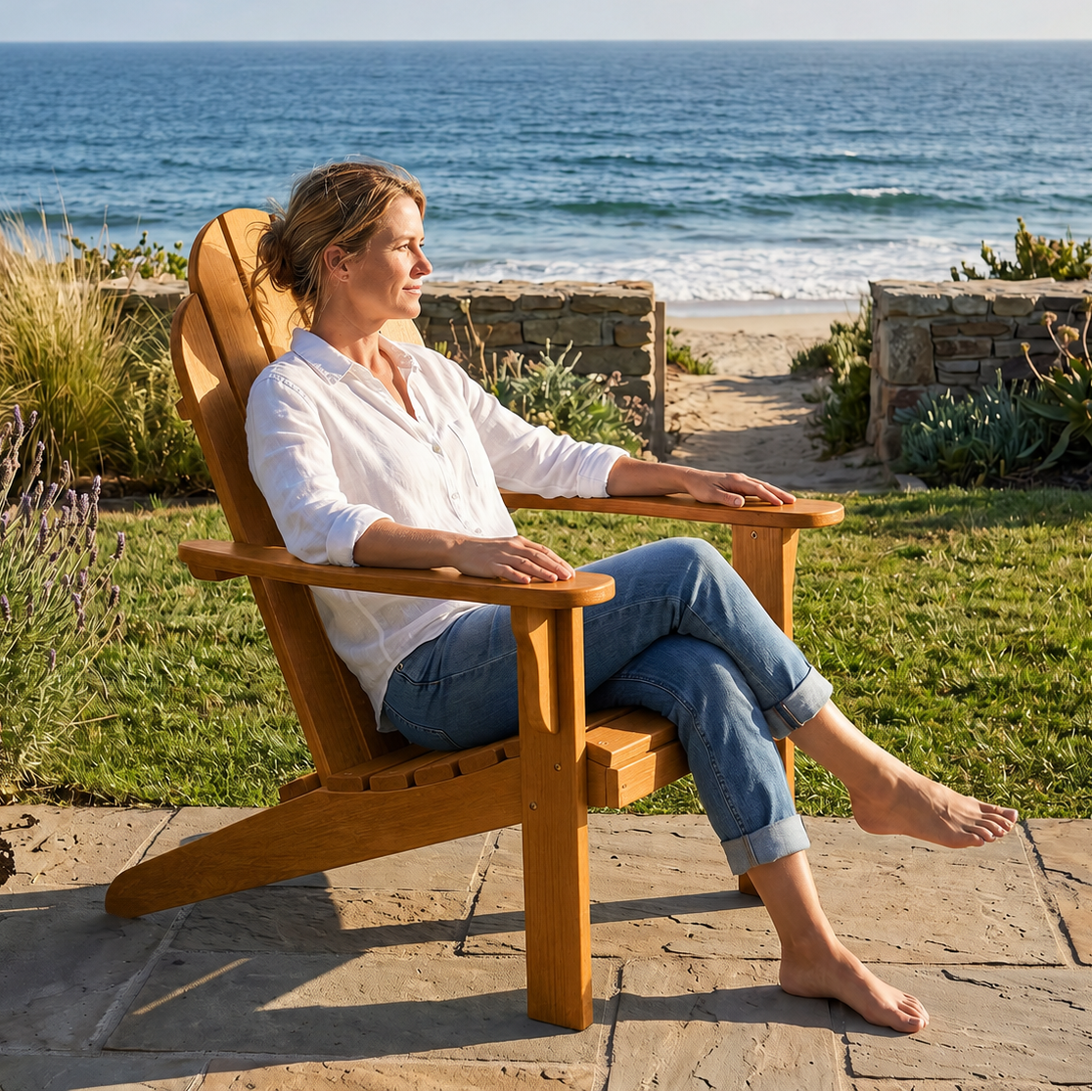 A woman sits barefoot in a wooden Adirondack-style outdoor chair on a stone patio, with grass, plants, and the ocean in the background.