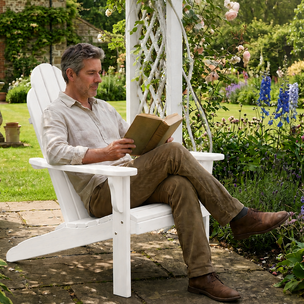 A man sits in a white wooden Adirondack-style garden chair on a stone patio, reading a book. The chair is under a white trellis arch decorated with pink roses, set in a lush, green garden with flowers and a stone house in the background.
