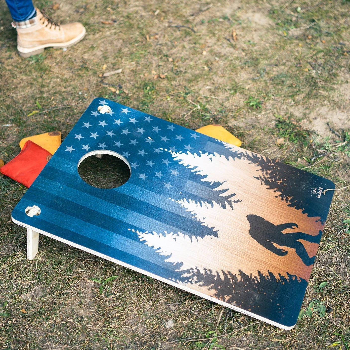 A cornhole board with a rustic American flag and forest design lies on grass outdoors. A person's legs and boots are visible at the top left, and red and yellow bean bags sit behind the board.