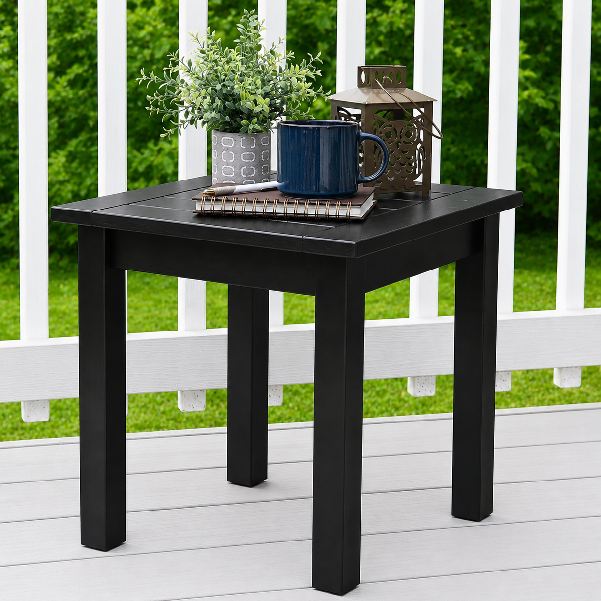 A square black outdoor side table with four legs, styled with a potted plant, a blue mug, a notebook, and a lantern, set on a white deck with a green garden background.