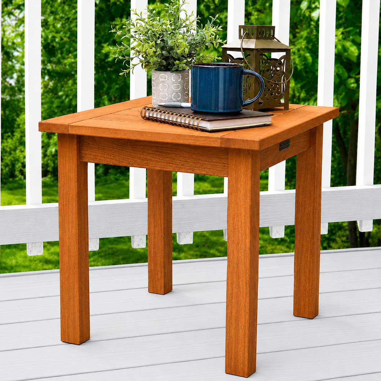 A square wooden end table with a warm natural oil finish, styled with a blue mug, notebook, potted plant, and lantern on a white deck with green foliage in the background.