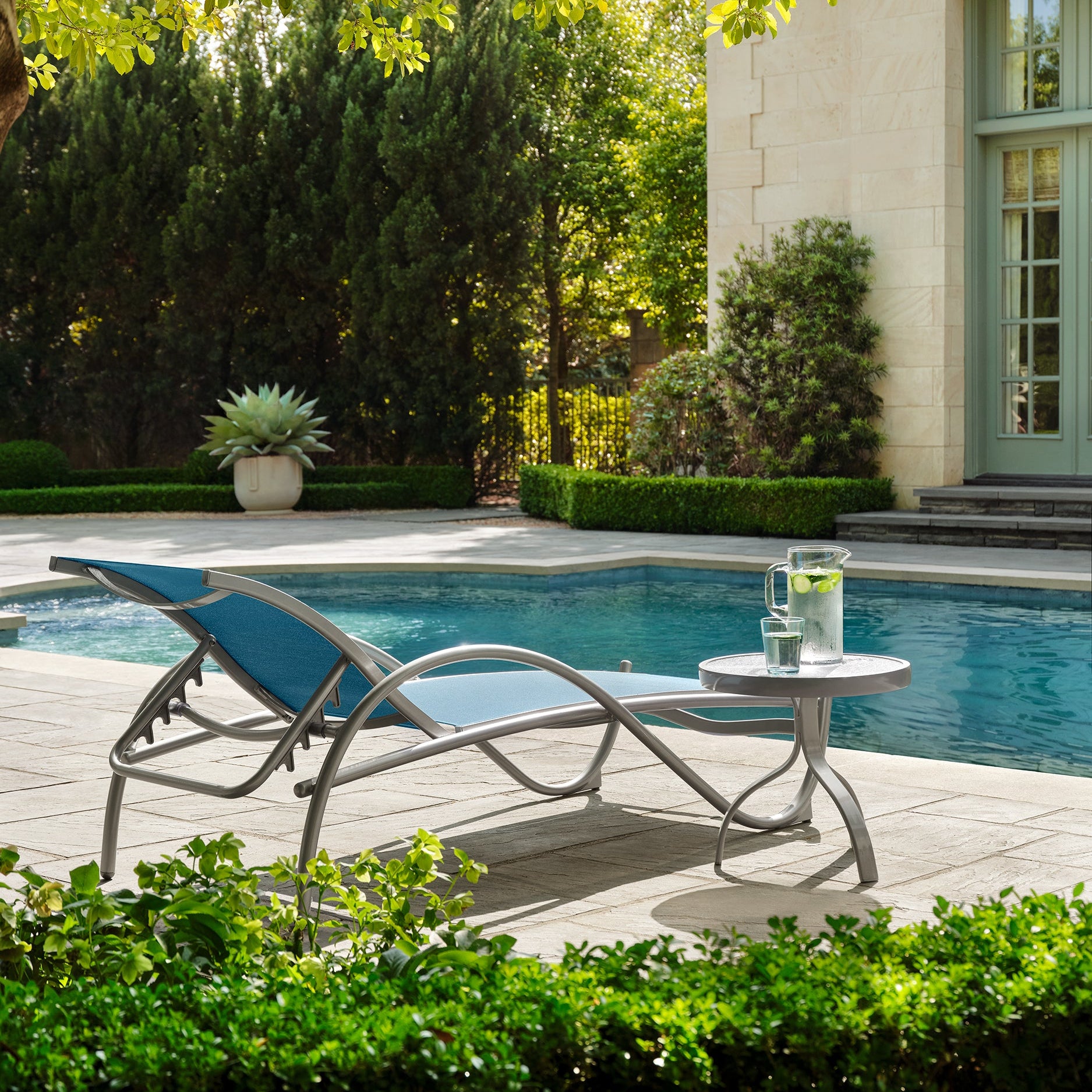 Chaise lounge by a pool with a table and glasses, surrounded by greenery.