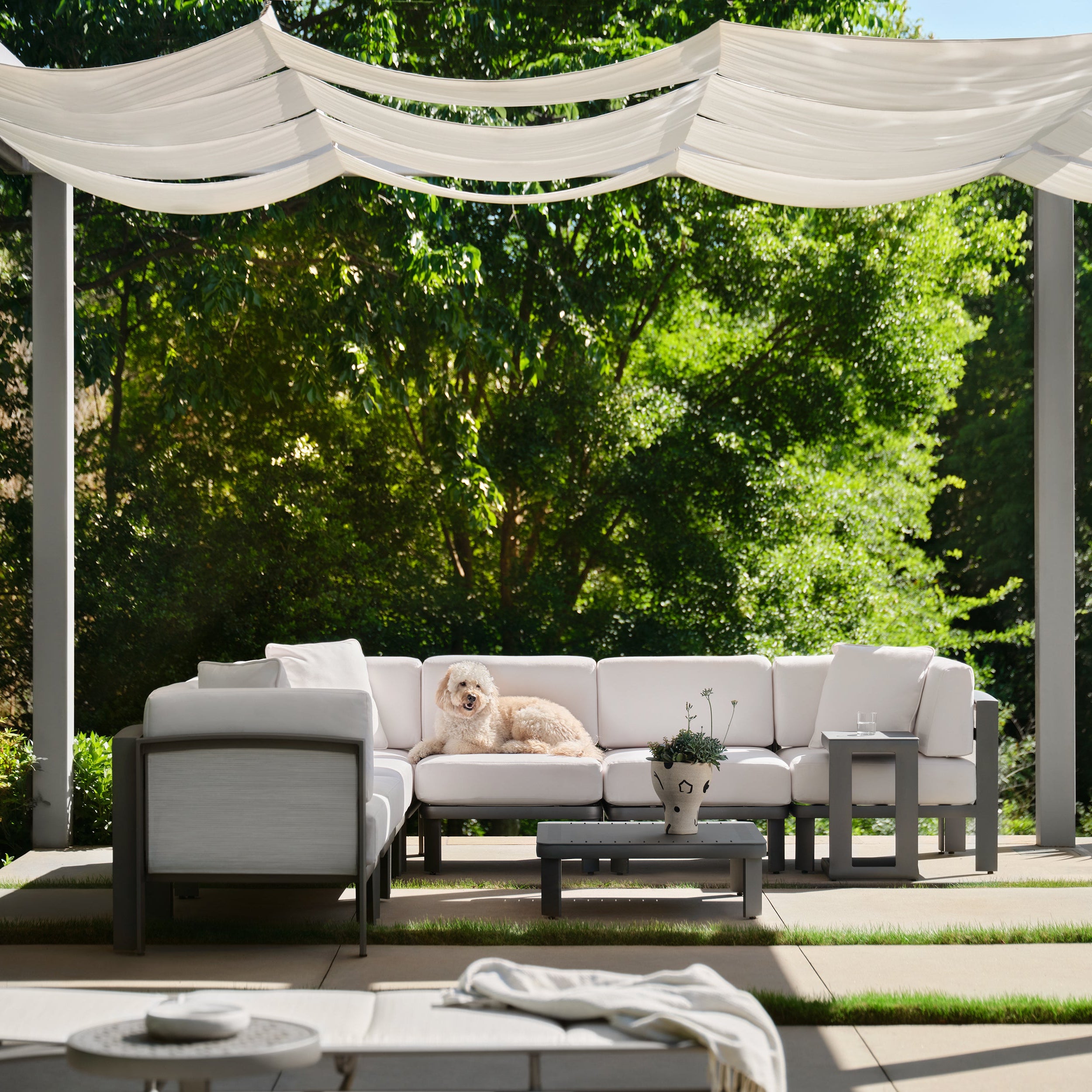 Outdoor patio with white sectional sofa, chairs, and a canopy under green trees.